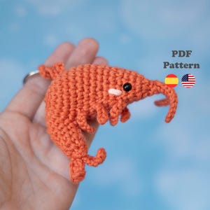 Puede incluir: Un llavero de camarón naranja tejido a ganchillo con ojos negros y un acento blanco. El camarón tiene un hocico curvado y patas pequeñas. El texto "PDF Pattern" es visible en la imagen, junto con las banderas de España y Estados Unidos.