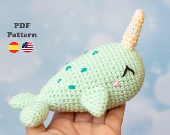 May include: A crocheted amigurumi narwhal toy with a light green body, a yellow and white horn, and black eyes. The narwhal is lying on its side with its tail curled up.