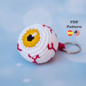 Puede incluir: Un llavero de ganchillo con forma de globo ocular, compuesto por una esfera blanca con venas rojas bordadas, un iris amarillo y una pupila negra. El texto PDF Pattern es visible junto a iconos de banderas española y estadounidense y una anilla.
