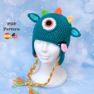 Puede incluir: Un gorro de monstruo de ganchillo turquesa con un ojo grande, cuernos pequeños y una lengua rosa. El gorro tiene orejeras y lazos trenzados con borlas naranjas y verdes. El texto "PDF Pattern" es visible.