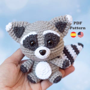 May include: A crocheted raccoon toy in grey, white, and black. The toy features large black eyes, a white chest, and a striped tail. The image includes the text "PDF Pattern" and flags for Spain and the United States.