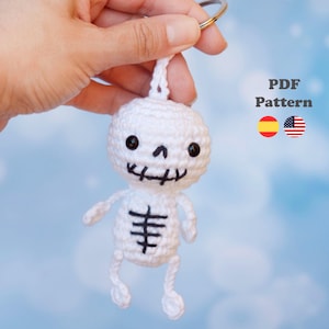 Puede incluir: Un llavero de esqueleto tejido a ganchillo en color blanco con ojos, nariz y boca bordados en negro, y una caja torácica bordada en negro en el torso. El texto PDF Pattern es visible junto a iconos de banderas española y estadounidense.