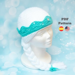 Può includere: Un cappello bianco all'uncinetto con un motivo a corona turchese e una lunga treccia bianca. Il cappello è esposto su una testa di manichino. Il testo "PDF Pattern" è visibile, insieme alle bandiere di Spagna e Stati Uniti.