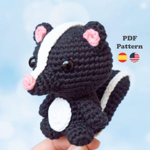 Può includere: Un giocattolo di puzzola all'uncinetto in bianco e nero con accenti rosa sulle orecchie e sul naso. La puzzola ha grandi occhi rotondi neri e una striscia bianca sulla schiena. Il testo "PDF Pattern" è visibile.