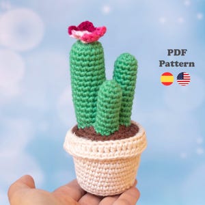 Puede incluir: Un cactus de ganchillo con tres tallos verdes en una maceta beige, con una pequeña flor rosa y blanca en el tallo más alto. El texto PDF Pattern es visible junto a iconos de banderas española y estadounidense, indicando un documento digital.