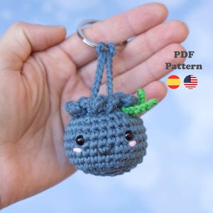 May include: A handmade, crocheted blueberry keychain with a smiling face, pink cheeks, and green leaves. The blueberry is attached to a silver keyring. The text "PDF Pattern" is visible in the image.