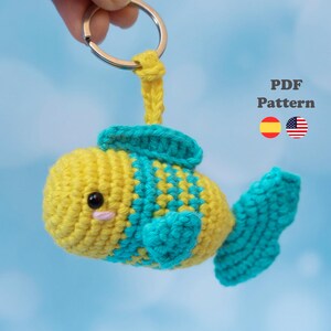 Puede incluir: Un llavero de pez tejido a mano en amarillo y turquesa. El pez tiene ojos negros y boca rosa. El llavero está sujeto a un anillo de metal plateado. La imagen también incluye el texto "PDF Pattern".