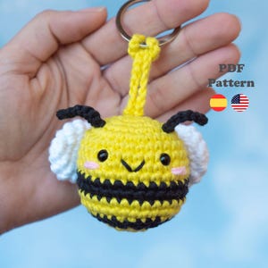 Puede incluir: Un llavero de abeja tejido a ganchillo con cuerpo redondo amarillo, rayas negras, alas blancas, antenas negras y mejillas rosadas. El texto PDF Pattern es visible junto a iconos de banderas española y estadounidense en una anilla de metal.