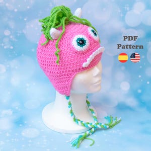 Peut inclure: Un chapeau rose crocheté avec un motif de visage de monstre, avec de grands yeux bordés de blanc avec des centres turquoise, des cornes blanches et des cheveux verts. Le chapeau a une mentonnière tressée avec des accents verts et bleus. Le texte "PDF Pattern" est visible.