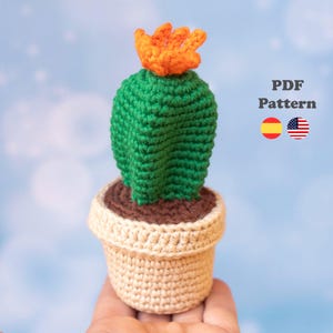 Puede incluir: Un cactus de ganchillo con un cuerpo verde brillante y una flor naranja en la parte superior, plantado en una maceta beige con una base de tierra marrón. El texto PDF Pattern es visible junto a iconos de banderas española y estadounidense.