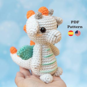 Puede incluir: Un juguete de dragón de ganchillo en blanco, naranja y verde azulado. El dragón tiene grandes ojos negros y sostiene un patrón PDF. El dragón tiene púas naranjas en la cabeza y la espalda, y alas verde azulado. El texto "PDF Pattern" es visible.
