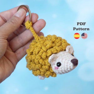 Puede incluir: Un llavero de erizo de ganchillo con un lomo texturizado de color amarillo mostaza y una cara de color crema con ojos negros, nariz marrón y mejillas rosadas. El texto "PDF Pattern" aparece junto a iconos de banderas.