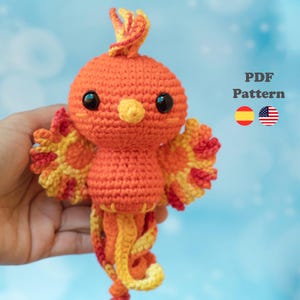 Puede incluir: Un juguete amigurumi de fénix tejido a ganchillo en tonos naranja, amarillo y rojo con ojos de seguridad negros y un pequeño pico amarillo. El texto PDF Pattern es visible a la derecha, junto a un icono de bandera española y estadounidense.