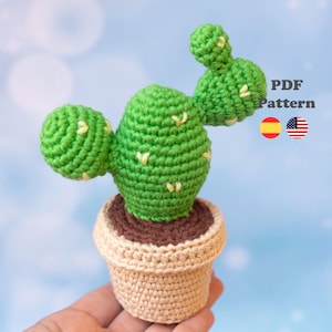 Puede incluir: Un cactus de ganchillo en una maceta beige con tierra marrón. El cactus es de color verde brillante con pequeñas puntadas bordadas en amarillo y tiene tres brazos redondeados. El texto a la derecha dice PDF Pattern con banderas.