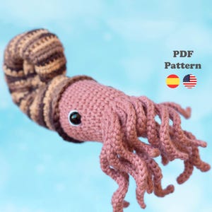 May include: A crocheted, pink and brown ammonite with black eyes and multiple tentacles. The image includes the text "PDF Pattern" and flags of Spain and the United States.