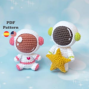 May include: Two crocheted astronaut dolls. One doll is seated, wearing a white spacesuit with pink accents and a pink heart. The other doll is holding a yellow star. Both dolls have white helmets and brown faces. The text "PDF Pattern" is visible.