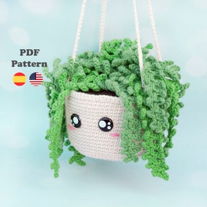 Puede incluir: Una planta colgante de crochet con una maceta blanca y hojas verdes. La maceta tiene una carita linda con ojos negros y mejillas rosadas. La planta está colgada de una cuerda blanca.