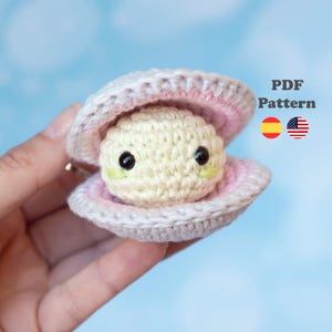 Puede incluir: Una perla de ganchillo en una concha. La perla es de color blanquecino con ojos negros y mejillas verdes. La concha es rosa claro por dentro y gris claro por fuera. El texto "PDF Pattern" es visible.