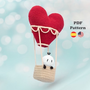 Puede incluir: Un globo aerostático de corazón tejido a crochet con una pequeña figura tejida a crochet en su interior. El globo es rojo y la figura es blanca. El globo tiene una cesta beige y una cuerda blanca atada a la parte superior. El texto "PDF Pattern" está en la esquina superior derecha de la imagen. Las banderas de España y Estados Unidos están debajo del texto.