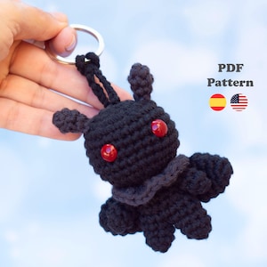 Puede incluir: Un llavero de ganchillo negro con forma de pequeña criatura con dos ojos de cuentas rojas, un cuello con volantes y dos orejas pequeñas. El artículo está unido a una anilla de metal plateado. El texto PDF Pattern aparece con dos banderas.