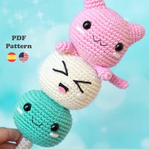 Może przedstawiać: Zestaw pluszowych zabawek amigurumi wykonanych na szydełku, przedstawiający trzy okrągłe postacie z różnymi wyrażeniami twarzy. Postać na górze to różowy kot z czarnymi oczami i białym otworem gęby. Postać w środku jest biała z czarnymi oczami i czarnym otworem gęby. Postać na dole jest turkusowa z czarnymi oczami i białym otworem gęby. Postacie są ułożone jedna na drugiej, przypominając japońskie ciasto mochi.