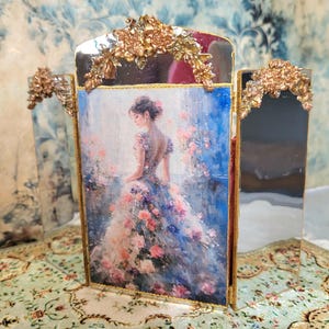 May include: A three-panel folding mirror featuring a central print of a person in a floral gown. The frame is edged in gold metal with ornate gold floral embellishments at the top of each panel. The side panels are reflective glass mirrors.