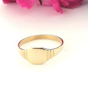 May include: A gold signet ring with a smooth, flat top. The ring has a simple, classic design.