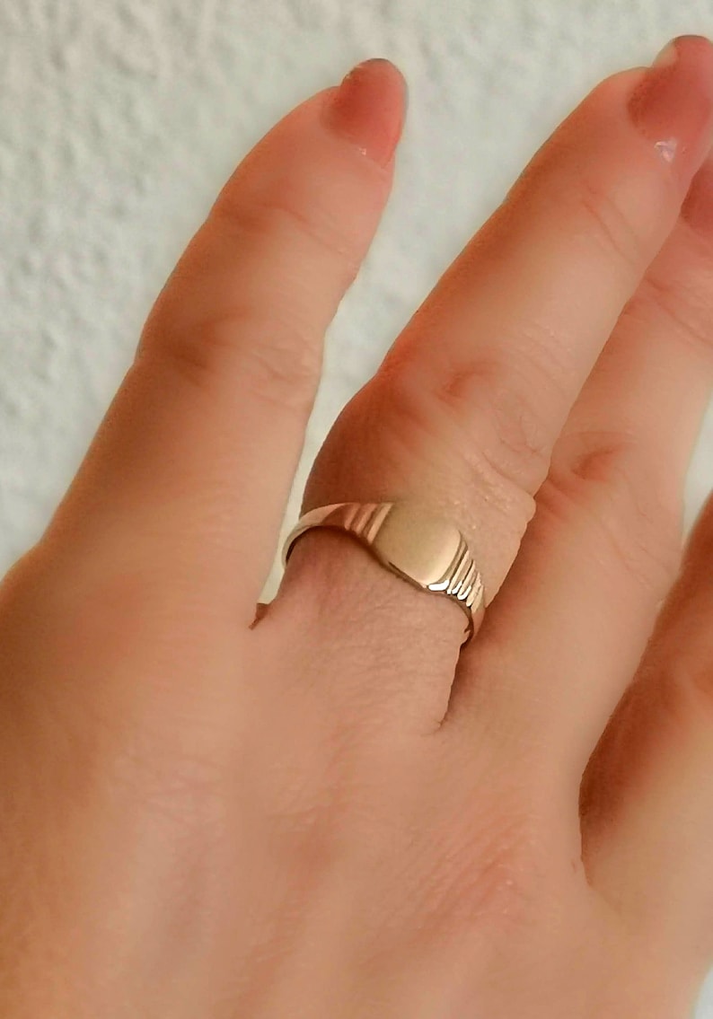 May include: A gold signet ring with a smooth, oval-shaped top. The ring is worn on a finger.