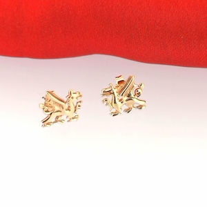 May include: A pair of gold-colored dragon stud earrings. The earrings are detailed with wings, claws, and a textured body. The earrings are displayed on a white surface with a red fabric in the background.