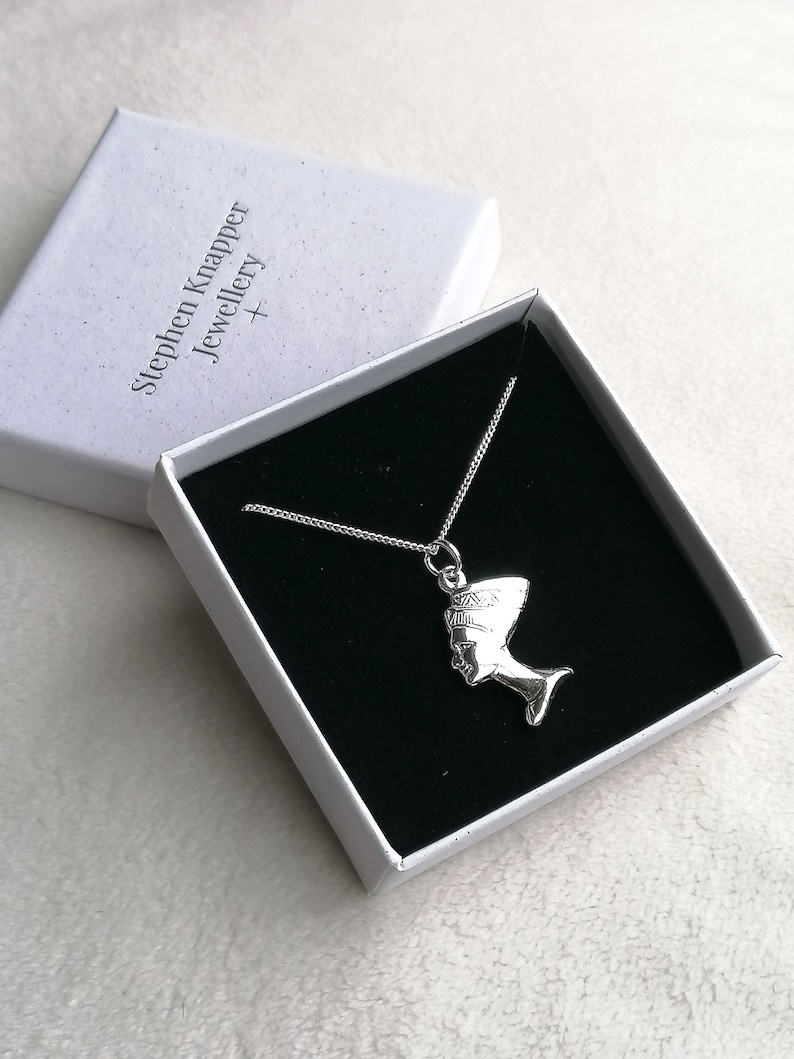 May include: A silver necklace with a pendant shaped like the profile of an Egyptian queen wearing a headdress. The necklace is in a white box with the text "Stephen Knapper Jewellery +" printed on the lid.