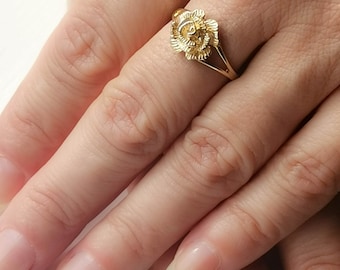 Può includere: Un anello dorato con un design a forma di rosa. L'anello è indossato su un dito.
