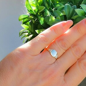 May include: A silver signet ring with a smooth, oval-shaped top. The ring is worn on a person's finger.