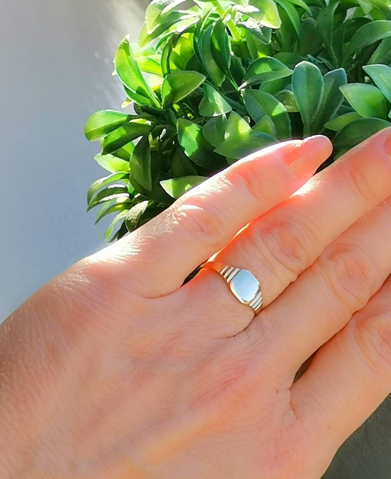 May include: A silver signet ring with a smooth, oval-shaped top. The ring is worn on a person's finger.