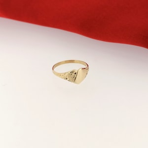 May include: A gold signet ring with a heart-shaped top. The ring has a detailed design on the band.