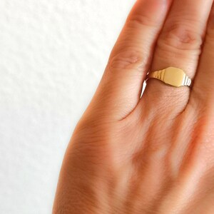 May include: A gold signet ring with a smooth, flat top. The ring is worn on a finger.