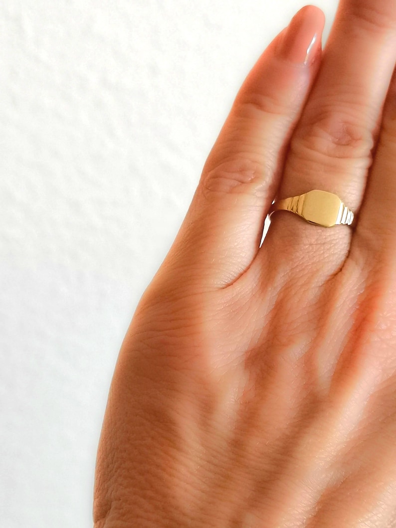 May include: A gold signet ring with a smooth, flat top. The ring is worn on a finger.