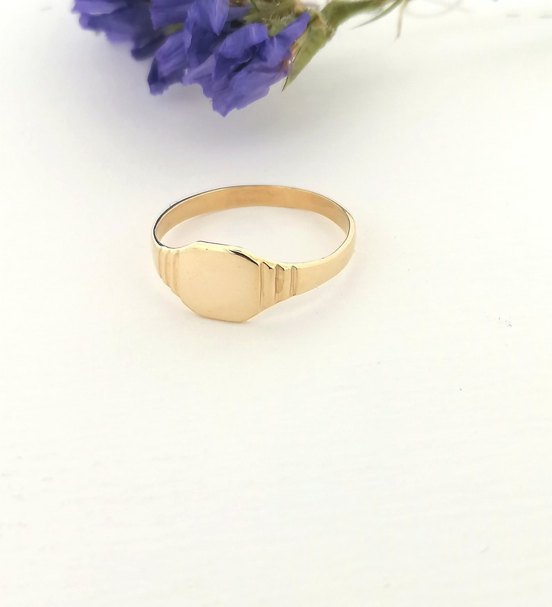 May include: A gold signet ring with a smooth, oval-shaped top. The ring is on a white surface.