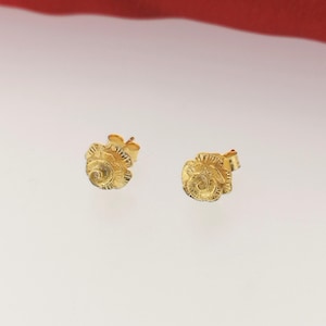 May include: Two gold-colored stud earrings. Each earring features a detailed, textured design resembling a small, stylized flower or shell. The earrings are set against a white background with a red backdrop.