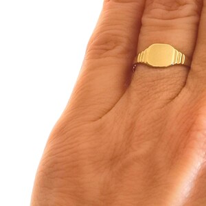 May include: A gold signet ring with a smooth, flat top. The ring is worn on a finger.