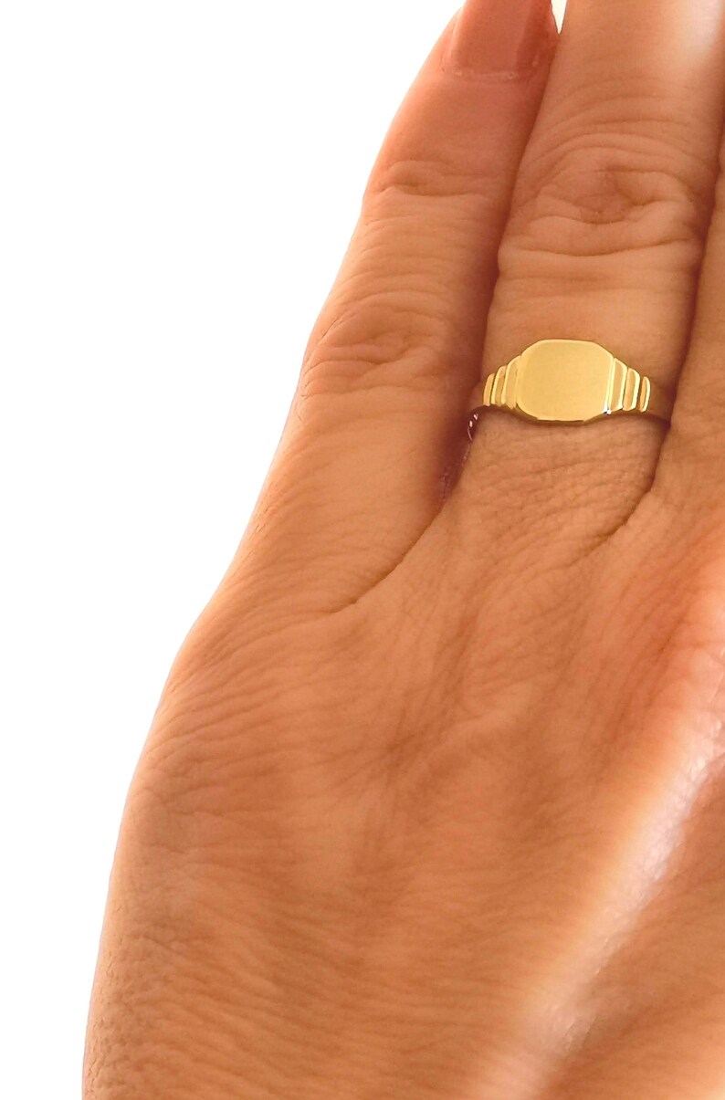 May include: A gold signet ring with a smooth, flat top. The ring is worn on a finger.