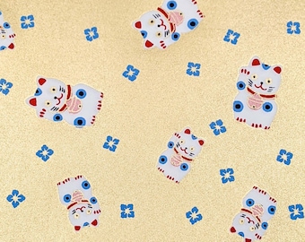 May include: A gold background with a repeating pattern of white lucky cats with blue eyes and red accents. The cats are holding a gold coin in their right paw and have a blue flower pattern on their chest. The background is covered in small blue flowers.