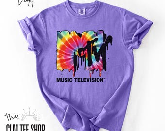 May include: A violet short-sleeve t-shirt featuring a large MTV logo graphic on the chest. The logo has a colorful tie-dye pattern with black paint-like drips. Below the logo, the text reads MUSIC TELEVISION in bold black capital letters.