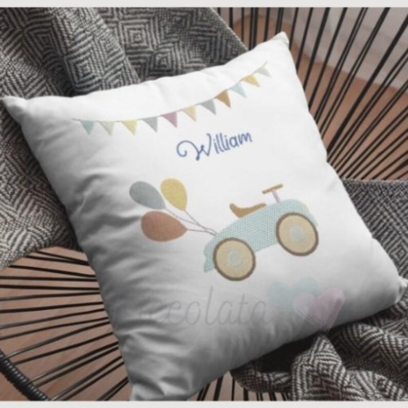 May include: White pillow with a blue and yellow toy car, three balloons, and the name "William" embroidered on it. The pillow has a string of colorful flags across the top.