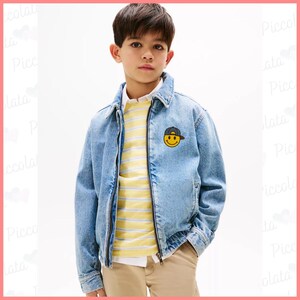 May include: A young person wearing a light blue denim jacket with a yellow and white striped shirt underneath. The jacket has a smiling face with a hat patch on the left side of the chest.