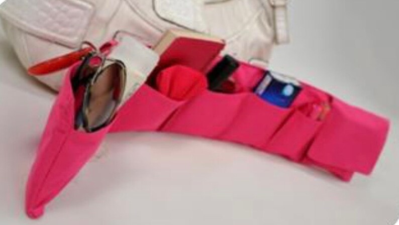 Pu&ograve; includere: Organizzatore in tessuto rosa con pi&ugrave; tasche per una borsa a mano. L'organizzatore ha una tasca per gli occhiali da sole, una tasca per un piccolo taccuino, una tasca per un telefono e una tasca per una bottiglia piccola.