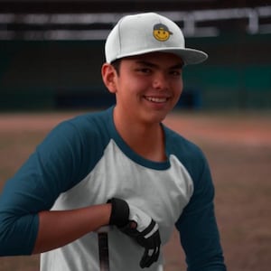 May include: A person wearing a white baseball cap with a yellow smiley face logo, a teal and white baseball jersey, and a black glove.