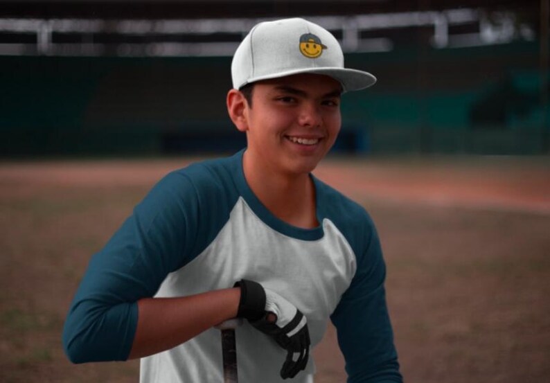 May include: A person wearing a white baseball cap with a yellow smiley face logo, a teal and white baseball jersey, and a black glove.