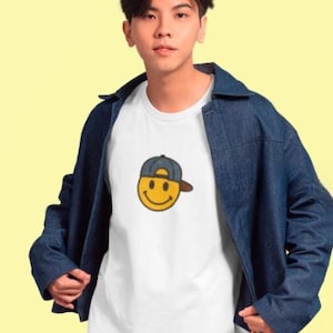May include: A white t-shirt with a yellow smiley face wearing a blue baseball cap. The person is wearing a blue denim jacket.