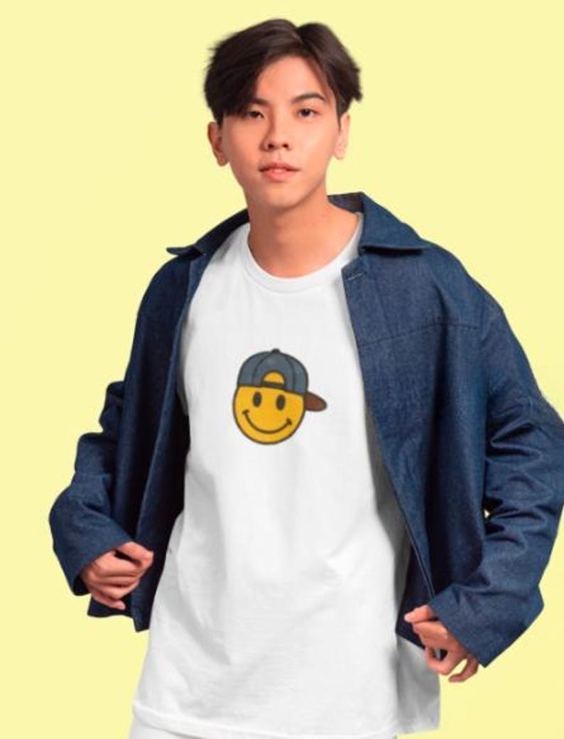 May include: A white t-shirt with a yellow smiley face wearing a blue baseball cap. The person is wearing a blue denim jacket.