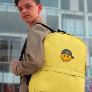 May include: A yellow backpack with a smiling face wearing a baseball cap. The backpack has a front pocket and adjustable straps.